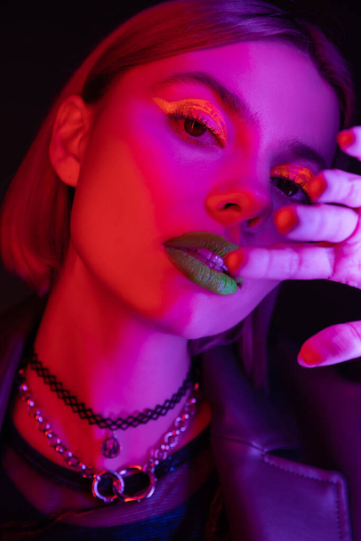 portret van jonge vrouw met heldere neon make-up hand in de buurt gezicht in paars licht op zwarte achtergrond - Foto, afbeelding