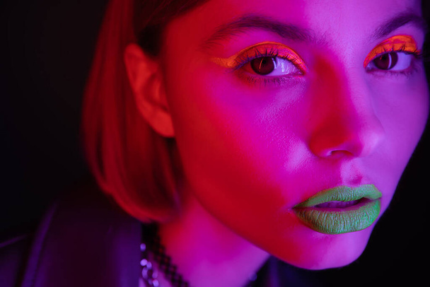 close up portrait of woman with glowing neon makeup in purple light on black background - Photo, Image