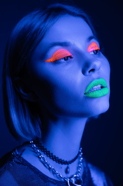 retrato de mulher elegante em maquiagem de néon colorido e colares olhando para longe no fundo azul escuro - Foto, Imagem
