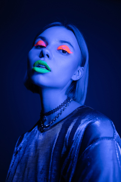 modna kobieta w błyszczącej bluzce i świecący neon makijaż patrząc na aparat fotograficzny na ciemnoniebieskim tle - Zdjęcie, obraz