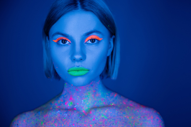 портрет женщины в ярком неоновом макияже и флуоресцентной краске тела, смотрящей на камеру, изолированную на темно-синем - Фото, изображение