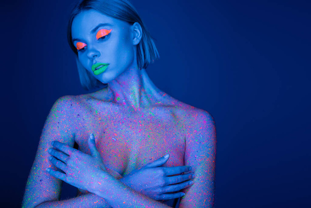 naked woman in neon makeup and bright paint splashes covering bust with hands isolated on dark blue - Foto, immagini