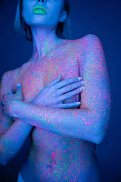 gedeeltelijk zicht op naakte vrouw met groene neonlippen en kleurrijke lichaamsverf die de borst bedekken met handen geïsoleerd op donkerblauw - Foto, afbeelding