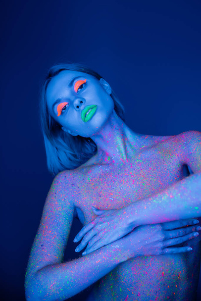 naked woman with vibrant makeup and neon paint on body isolated on dark blue - Photo, Image
