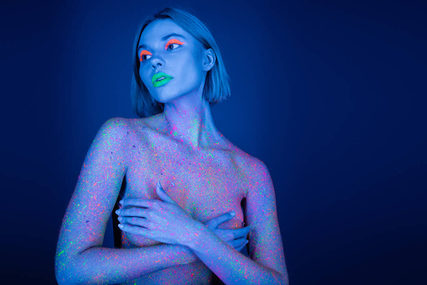 naked woman in neon makeup and bright paint splashes covering bust with hands and looking away isolated on dark blue - Photo, image