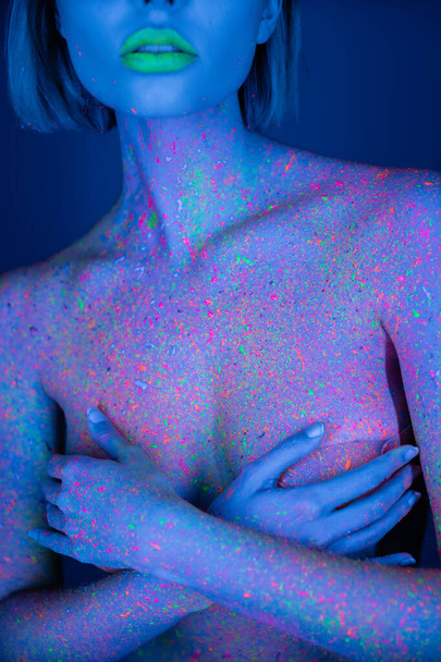 Ausgeschnittene Ansicht einer nackten Frau mit grünen Neon-Lippen und hellen Farbspritzern auf die Brust, die den Körper bedeckt, isoliert auf dunkelblau - Foto, Bild