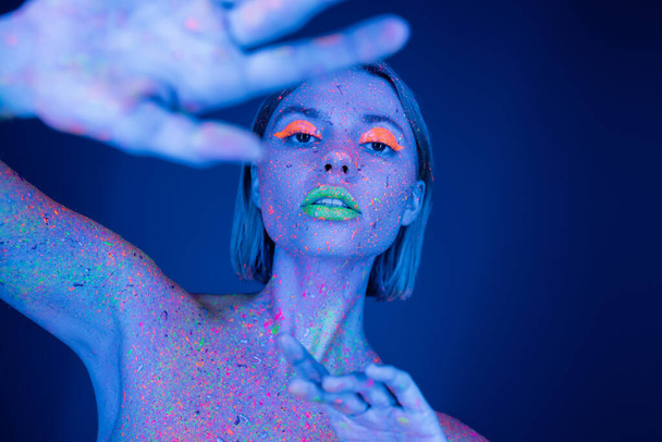 vrouw in gloeiende make-up en neon bodypaint poseren met wazig uitgestrekte hand geïsoleerd op donkerblauw - Foto, afbeelding