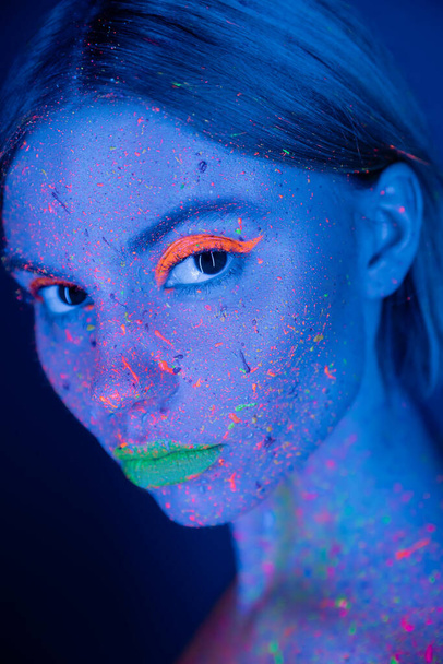 portret kobiety z neonowym makijażem i fluorescencyjną farbą na twarzy odizolowanej na ciemnoniebiesko - Zdjęcie, obraz
