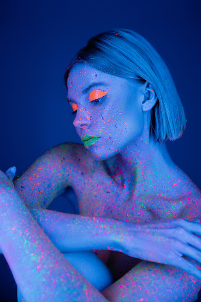 nude woman with neon makeup and body in fluorescent paint isolated on dark blue - Foto, Imagen