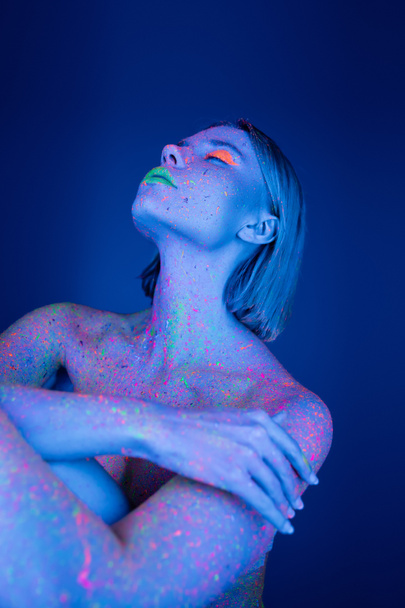 обнаженная женщина в ярком макияже и неоновой краске тела позирует на темно-синем фоне - Фото, изображение