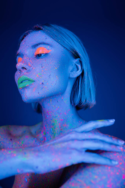 young woman with neon makeup touching body colored in fluorescent paint isolated on dark blue - Photo, Image