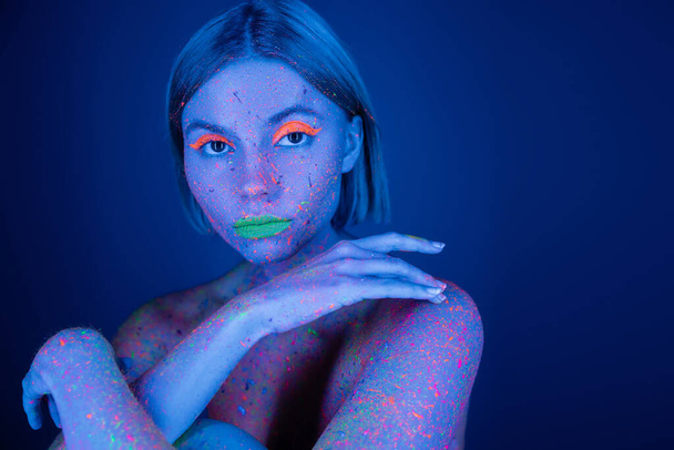 nude woman in vibrant body paint and neon makeup looking at camera isolated on dark blue - Foto, Bild