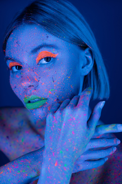žena se zářícím make-upem a barevnými skvrnami pózující v neonovém světle izolované na tmavě modré - Fotografie, Obrázek
