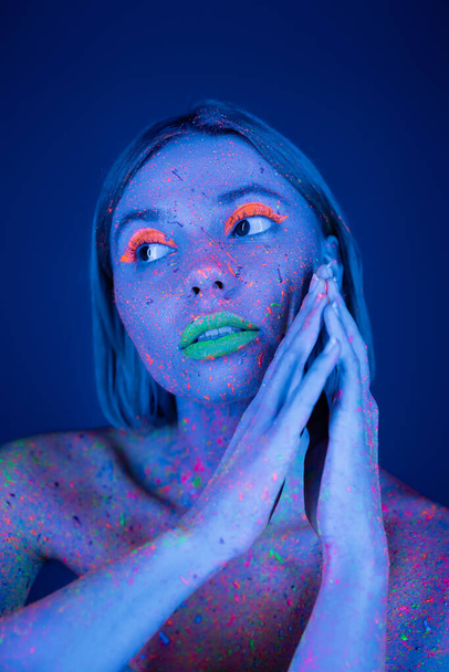 mujer joven con maquillaje de neón vibrante y salpicaduras de pintura brillante en el cuerpo mirando hacia otro lado aislado en azul oscuro - Foto, imagen