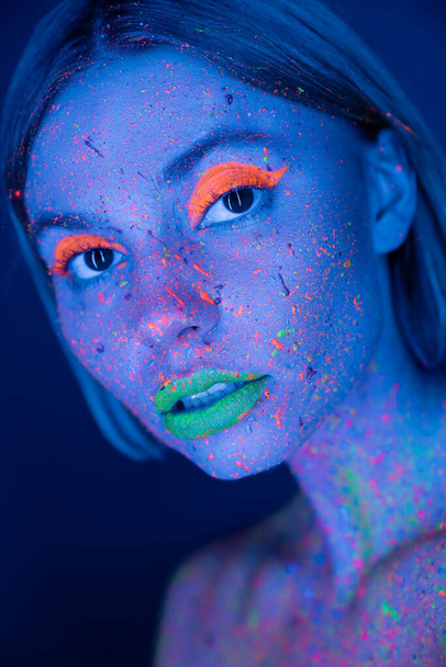 retrato de mujer con maquillaje brillante y pintura de neón en la cara aislada en azul oscuro - Foto, imagen