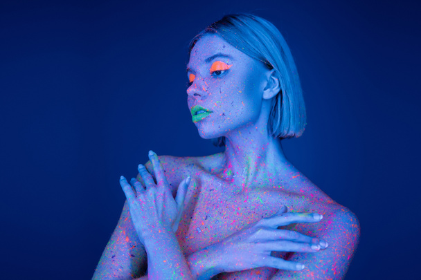 mulher nua em maquiagem de néon e respingos de tinta brilhante no corpo posando com braços cruzados isolados em azul - Foto, Imagem