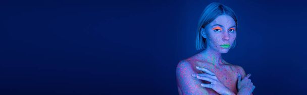 Frau in leuchtendem Neon-Make-up und bunten Farbspritzern posiert mit verschränkten Armen auf dunkelblauem Hintergrund, Banner - Foto, Bild