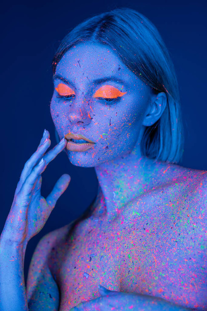 mulher sensual com maquiagem de néon e tinta brilhante no corpo tocando lábios isolados em azul escuro - Foto, Imagem