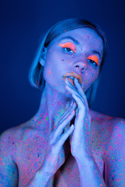 nude woman in glowing makeup and neon body paint holding hands near face isolated on dark blue - Photo, Image