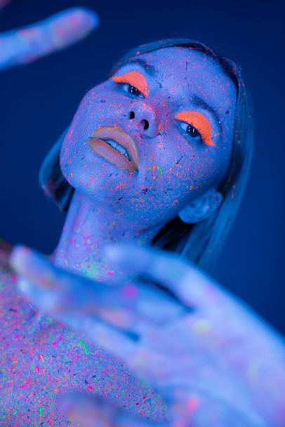 sensual woman with bright neon makeup and fluorescent paint posing on blurred foreground isolated on dark blue - Photo, Image