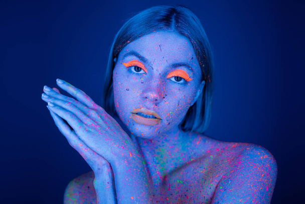 mooie vrouw in neon make-up en kleurrijke verf op zoek naar camera geïsoleerd op donkerblauw - Foto, afbeelding