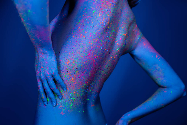 bijgesneden weergave van naakte vrouw in kleurrijke neon verf poseren geïsoleerd op donkerblauw - Foto, afbeelding