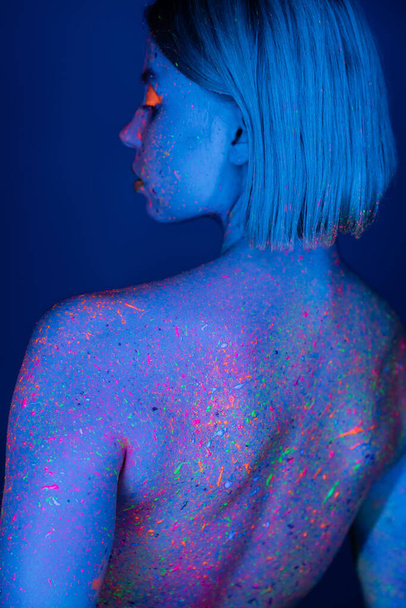 vista posterior de la mujer con salpicaduras de neón brillante en el cuerpo aislado en azul oscuro - Foto, Imagen