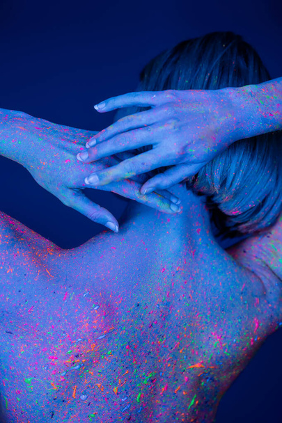濃い青で隔絶された頭の後ろに手でポーズをとった鮮やかなネオンボディペイントの女性の後ろ姿 - 写真・画像