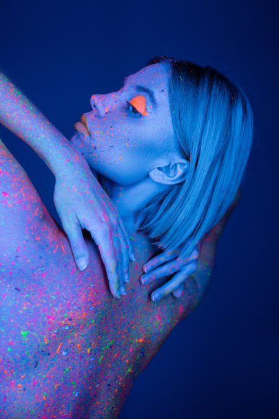 smyslná žena v barevné neonové barvy těla dotýkající se zpět izolované na tmavě modré - Fotografie, Obrázek