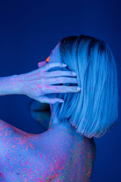 νεαρή γυναίκα χρωματισμένη με νέον χρώμα αγγίζοντας τα μαλλιά απομονώνονται σε σκούρο μπλε - Φωτογραφία, εικόνα