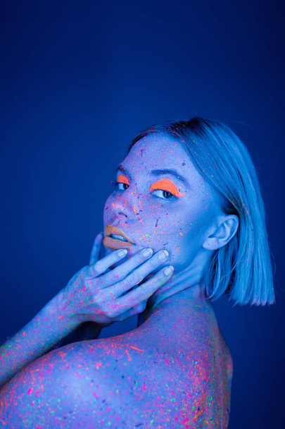 Canlı neon yüzlü genç bir kadın. Yüzüne dokunan renkli bir vücut. Koyu mavi renkli bir kameraya bakıyor. - Fotoğraf, Görsel