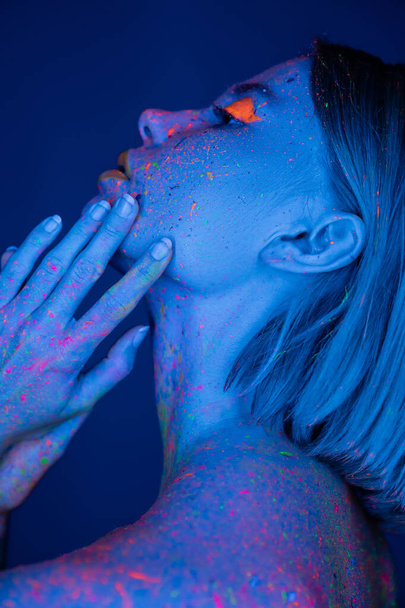 ネオンメイクの女性の側面図と濃い青で隔絶された顔の近くに手を持つボディペイント - 写真・画像