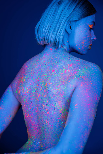 back view of woman with vibrant neon paint splashes on body isolated on dark blue - Photo, Image