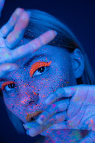 woman in neon makeup and bright paint splatters posing with blurred hands near face isolated on dark blue - Photo, Image