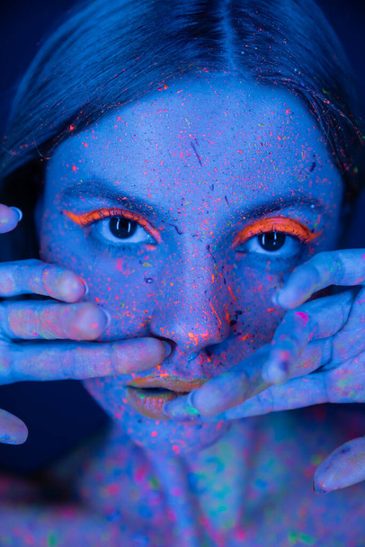 portrét ženy držící se za ruce poblíž obličeje s neonovým make-upem a fluorescenční barvou izolovanou na tmavě modré - Fotografie, Obrázek