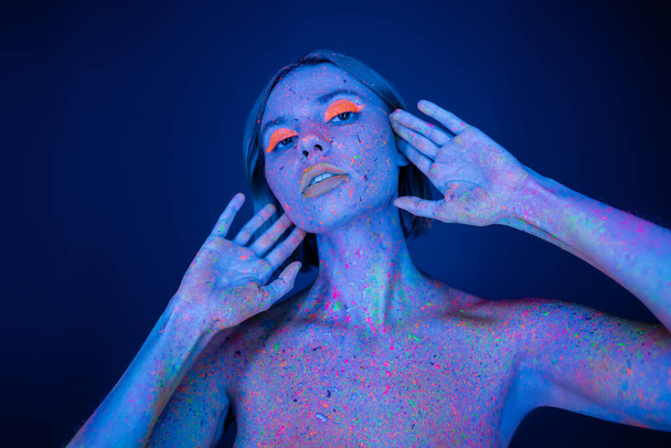 nackte Frau in Neon-Make-up und Körper in glühender Farbe posiert mit Händen in Gesichtsnähe isoliert auf dunkelblau - Foto, Bild