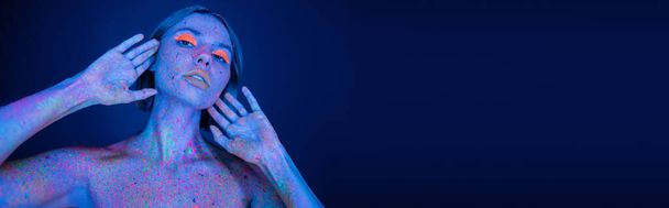 jonge vrouw met neon make-up en heldere verf spatten op het lichaam op zoek naar camera geïsoleerd op donkerblauw, banner - Foto, afbeelding