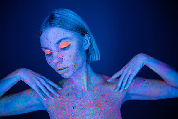 красивая женщина в ярком макияже и светящийся неоновой краски тела позирует изолированы на темно-синий - Фото, изображение