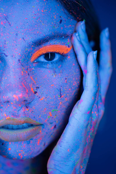 portrait en gros plan d'une femme recadrée touchant le visage avec du maquillage au néon et des éclaboussures de peinture vibrantes isolées sur du bleu foncé - Photo, image