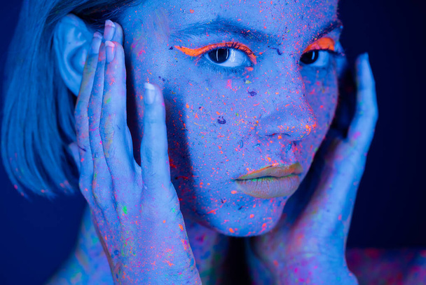 портрет женщины в неоновом макияже и флуоресцентной краске, держащей руки возле лица на темно-синем  - Фото, изображение