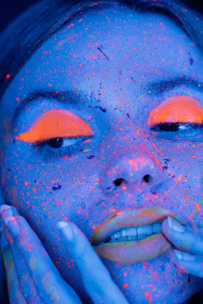 Nahaufnahme Porträt einer Frau mit fluoreszierendem Make-up, die ihr Gesicht in blauem Neonlicht berührt  - Foto, Bild