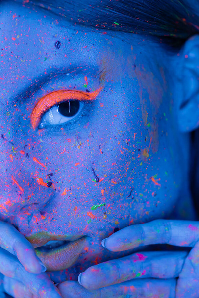 visão parcial da mulher de mãos dadas perto do rosto com maquiagem de néon e salpicos de tinta brilhante na luz azul  - Foto, Imagem