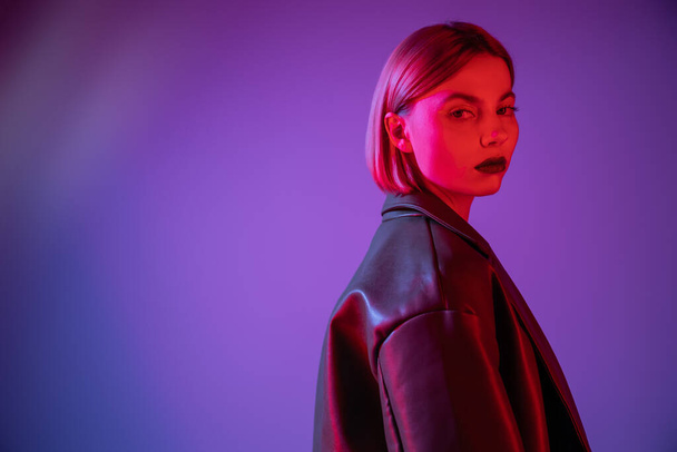 young and stylish woman looking at camera in red light on purple and blue background - Valokuva, kuva