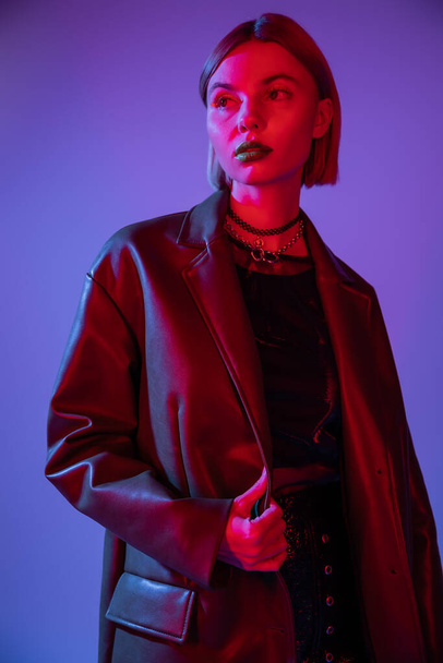 trendy woman in leather jacket and necklaces looking away in red light on blue background - Photo, Image