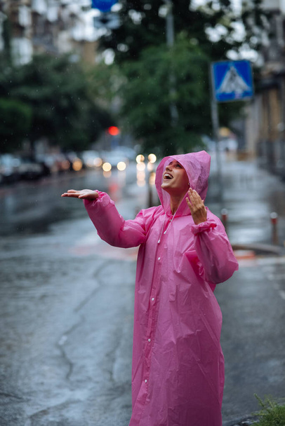 Young smiling woman with a pink raincoat on the street while enjoying a walk through the city on a rainy day. - Фото, изображение