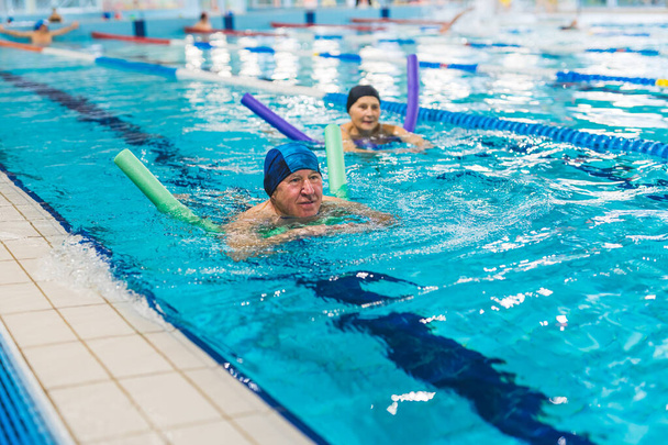 Активний позитивний кавказький одружений старший дорослий пара плаває в спортивному басейні. Люди в шапочках для плавання використовують барвисту локшину для пінного басейну. Високоякісна фотографія
 - Фото, зображення