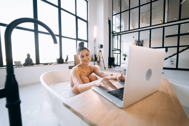 Νεαρή γυναίκα που εργάζεται σε φορητό υπολογιστή, ενώ λαμβάνει μια μπανιέρα στο σπίτι - Φωτογραφία, εικόνα