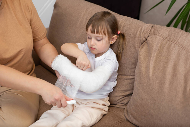 Mom wraps a bandage around her daughters broken arm. Gypsum. - Zdjęcie, obraz