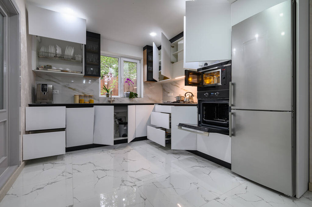 Uma cozinha deslumbrante com um design branco e moderno, um piso de mármore luxuoso e prateleiras de arrancar para facilitar o acesso. Fornos porta aberta. - Foto, Imagem