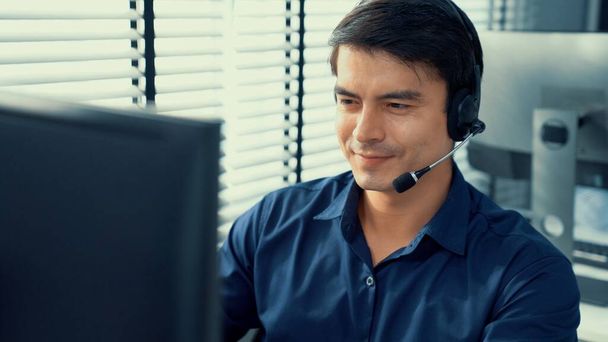 Young competent asian male call center agent working at his computer while simultaneously speaking with customers. Concept of an operator, customer service agent working in the office with headset. - Photo, Image